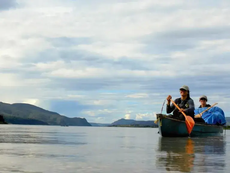 La imagen muestra a dos personas en una canoa en mar abierto con montañas al fondo.